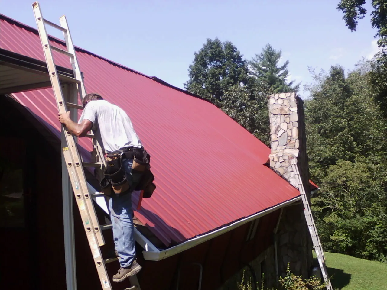 Roofer on a ladder beside a red metal roof and stone chimney — Soffit & Fascia Repair by Roofing Brothers in Laura